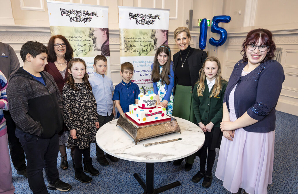 Photograph of HRH The Countess of Wessex, PSC Founder & CEO Ruth Pearse, and a group of children & young people surrounding a PSC birthday cake with a 15 balloon in the background