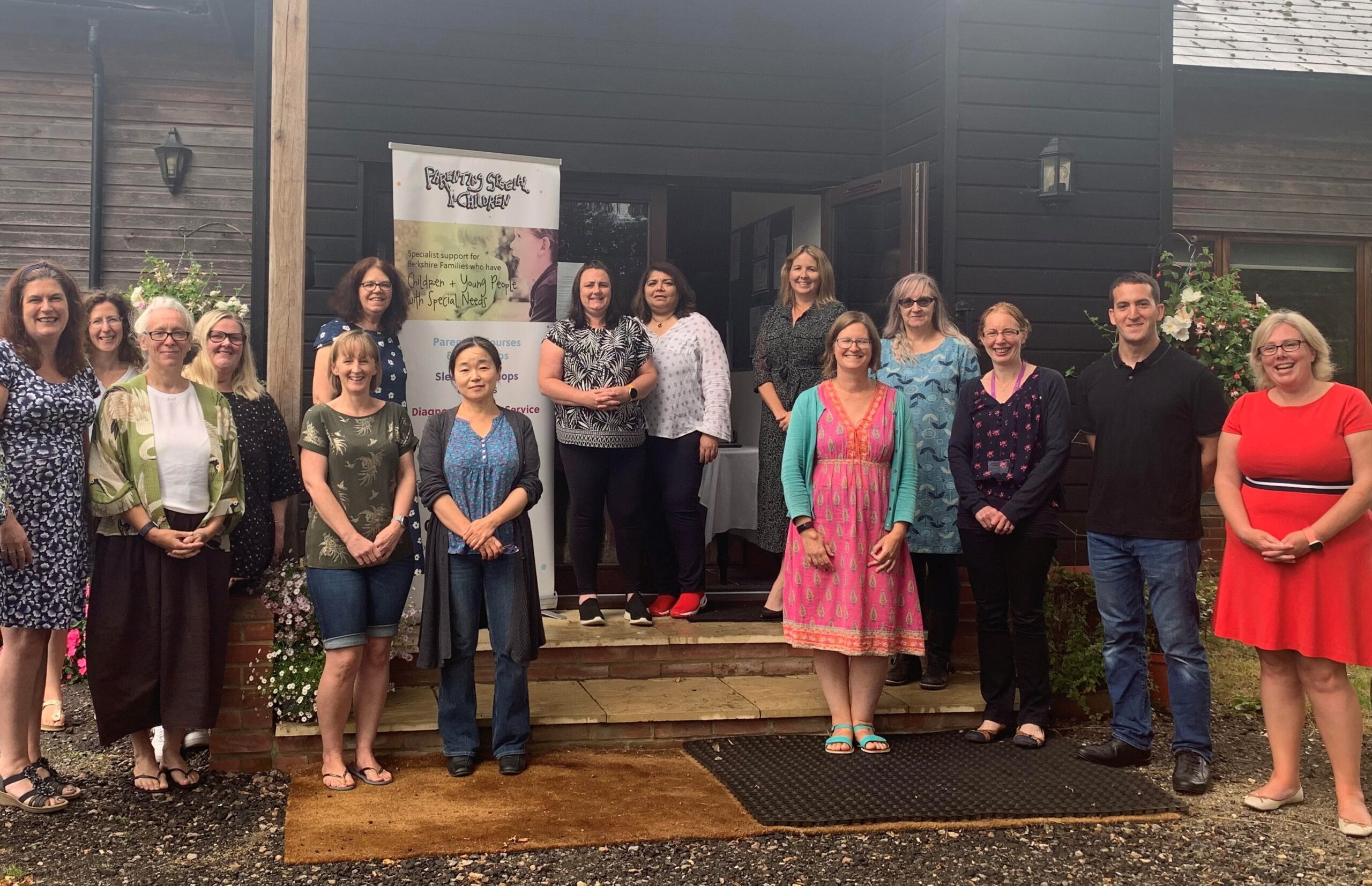 [A group photo showing 16 people standing in front of a building. They are standing on steps and a small porch, posing for the picture. There is a banner in the background that reads 'Parents Special Children' and mentions 'Specialist support for families who have children & young people with special needs.' The group consists of men and women, all casually dressed, and they are smiling at the camera. The setting appears to be an outdoor area with flowers and greenery around.]