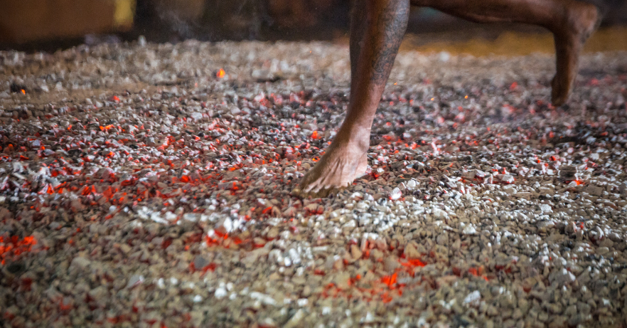 The image depicts a person walking or running barefoot over a bed of hot coals or embers, a practice commonly known as firewalking.