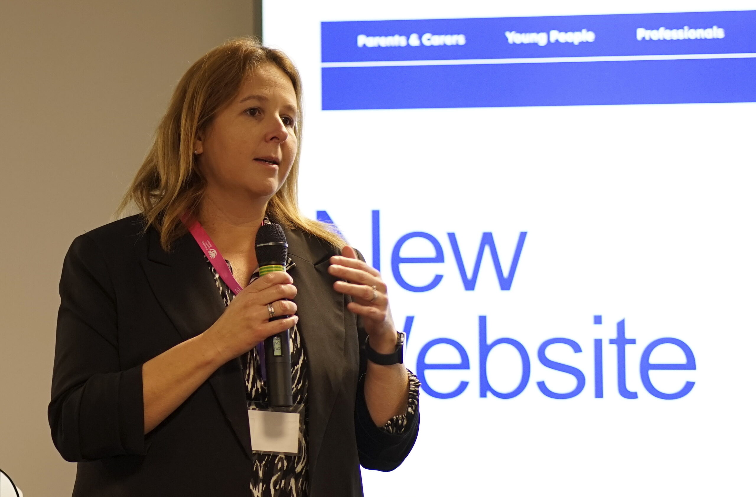 PSC Trustee Sarah Croxford stands speaking into a microphone, presenting in front of a screen displaying the text "New Website." The screen also has navigation options labeled "Parents & Carers," "Young People," and "Professionals." She wears a black blazer and a pink lanyard.