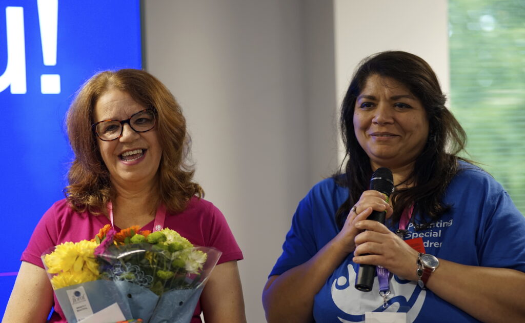 A close-up photo of two women (Ruth and Zobia) from the Parenting Special Children team at a presentation or event. The woman on the left (Ruth), wearing a bright pink T-shirt with the organization’s logo, is smiling and holding a bouquet of colorful flowers wrapped in blue paper. The woman on the right (Zobia), in a blue T-shirt with the same logo, is holding a microphone and smiling softly, as if addressing the audience. Behind them, a bright blue screen is visible, adding a cheerful backdrop to the scene.