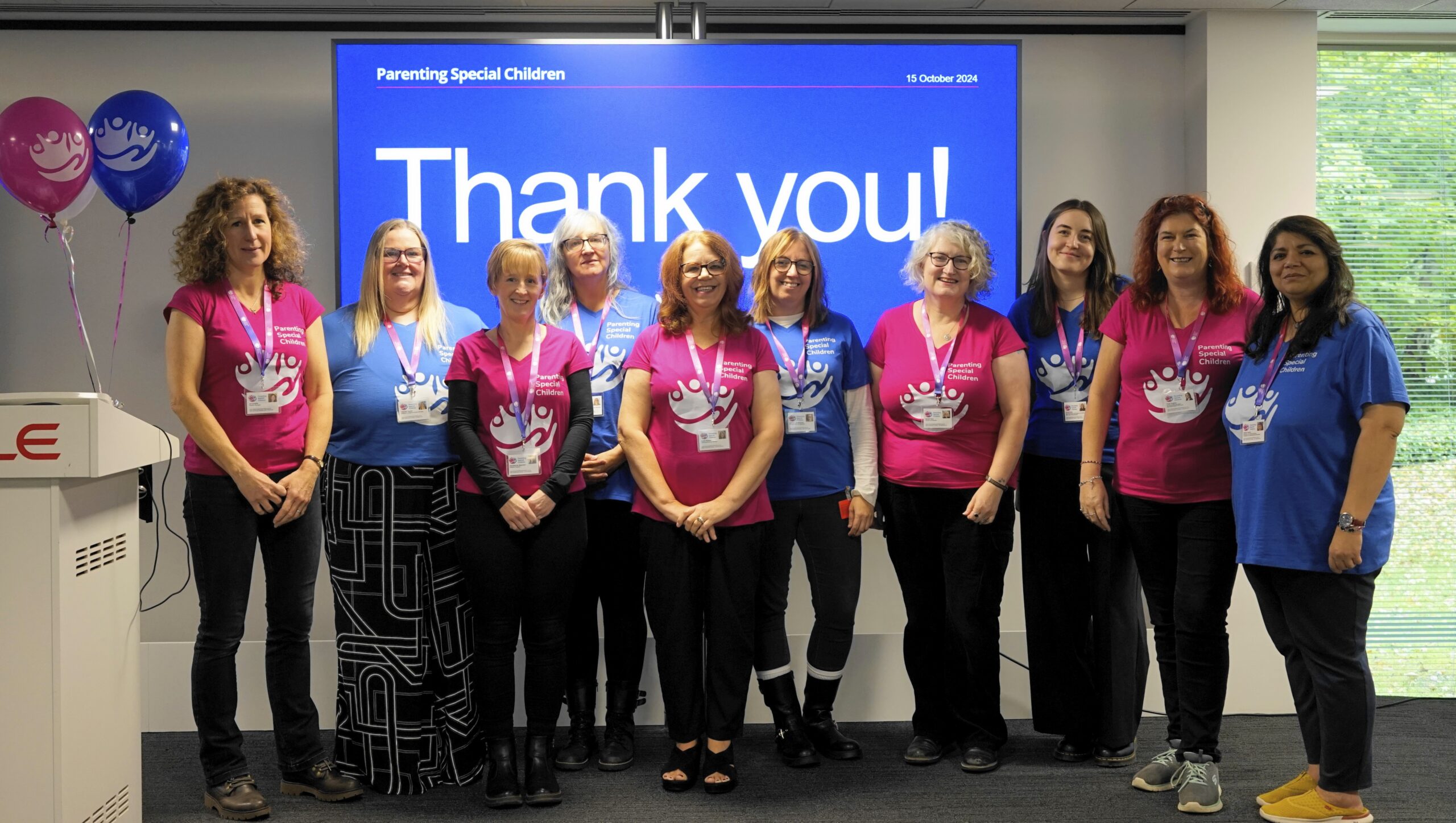 A group photo of the Parenting Special Children team, taken in front of a large screen that reads "Thank you!" dated October 15, 2024. The team members are standing in a row, smiling and wearing branded T-shirts in either bright pink or blue, each with the Parenting Special Children logo. The team members are wearing name badges and lanyards, suggesting a formal or professional event. Pink and blue balloons with the same logo are visible to the left of the group.