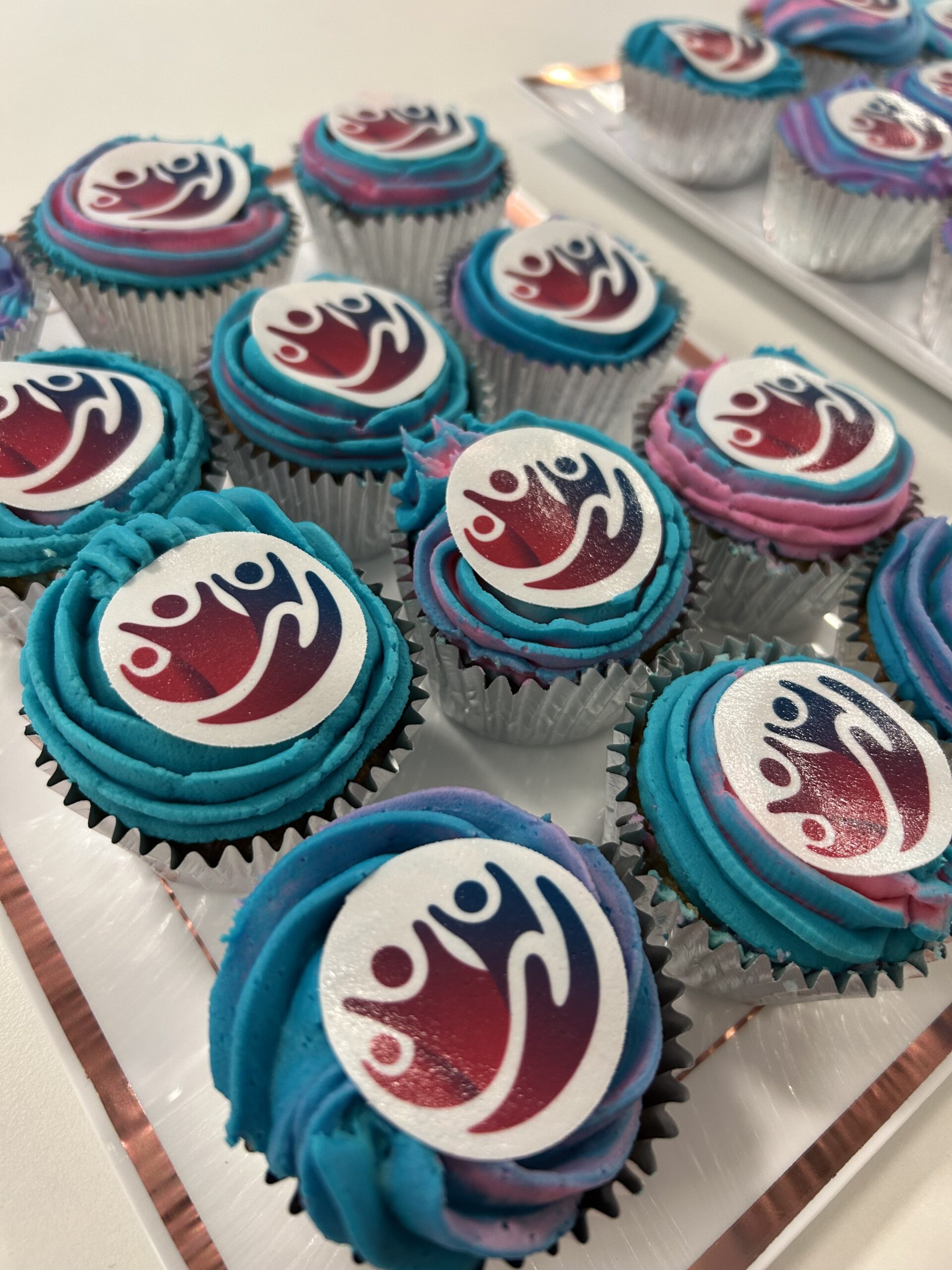 Cupcakes with blue and purple frosting, each topped with an edible PSC logo. The cupcakes are arranged on a tray with a white background.